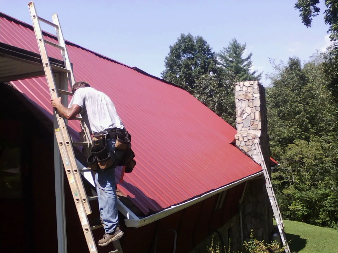 Roofer on a ladder beside a red metal roof and stone chimney — Soffit & Fascia Repair by Roofing Brothers in Leoma